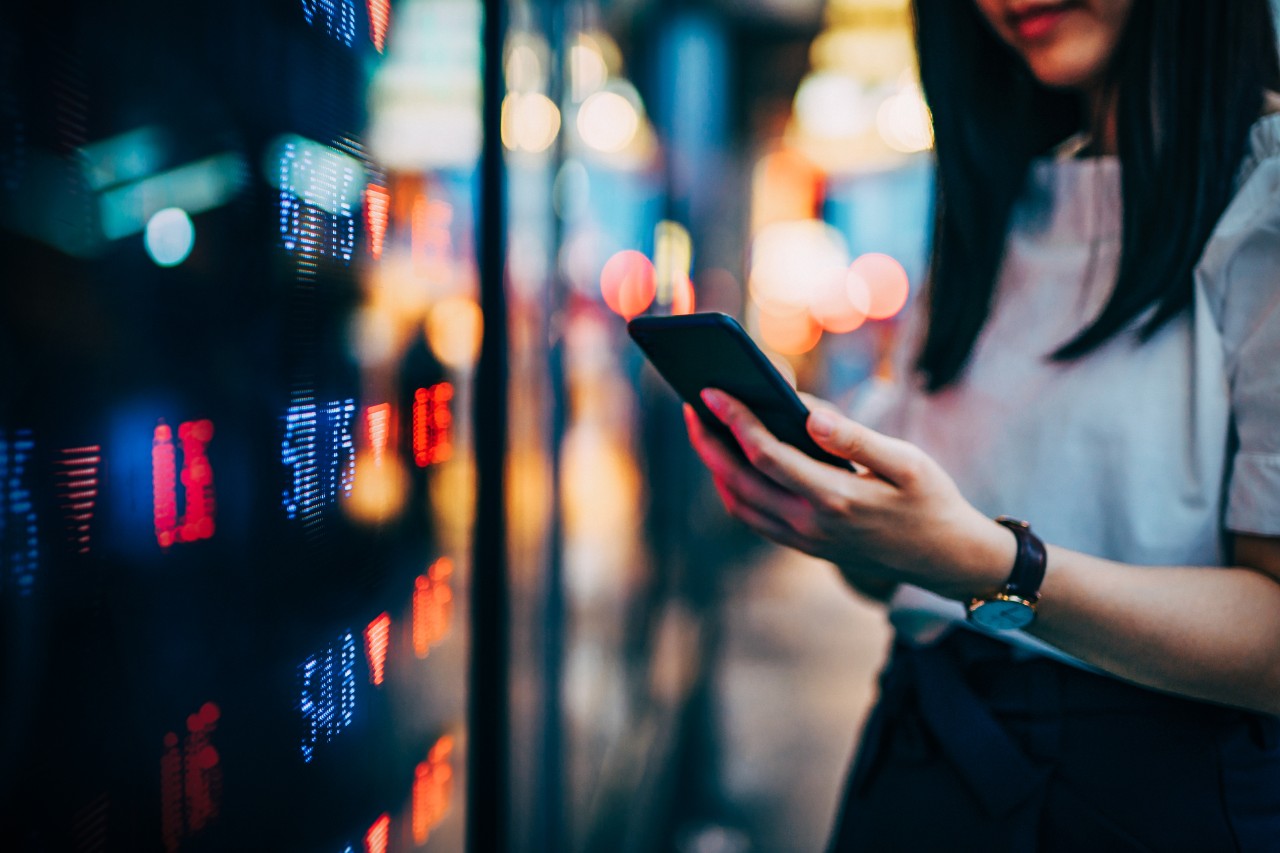 Businesswoman checking financial trading data on smartphone by the stock exchange market display screen board in downtown financial district showing stock market crash sell-off in red colour